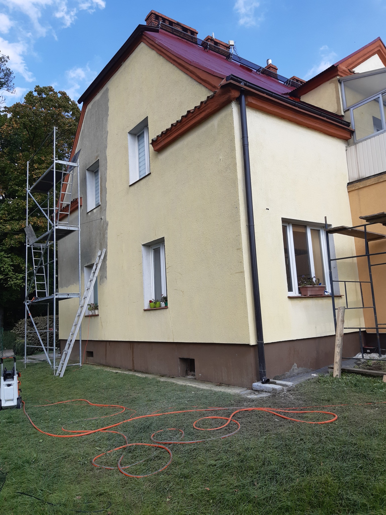 Dwukondygnacyjny dom z żółtą elewacją w trakcie czyszczenia; widoczne rusztowanie i myjka ciśnieniowa, fragment elewacji umyty, kontrastujące ze sobą powierzchnie, czerwona dachówka.