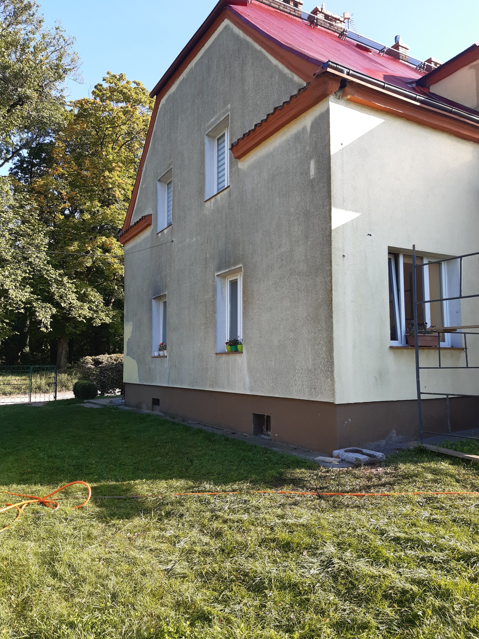 Elewacja domu z widocznymi śladami czyszczenia, czerwony dach, pomarańczowy wąż ogrodowy leżący na trawie.