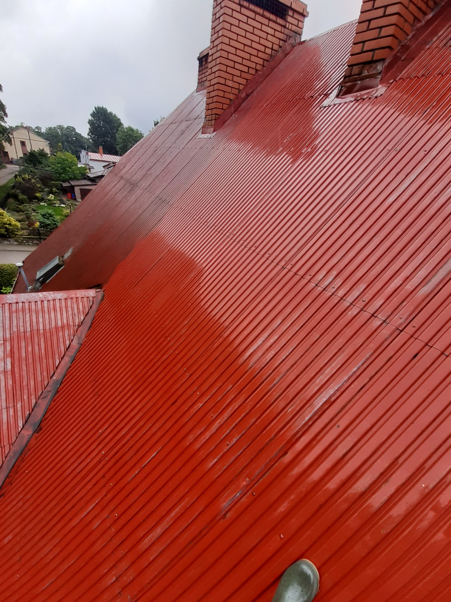 Czerwony, blaszany dach po umyciu, z widocznymi mokrymi śladami i ceglanym kominem w tle oraz fragmentem nogi w gumowym bucie w dolnym rogu.