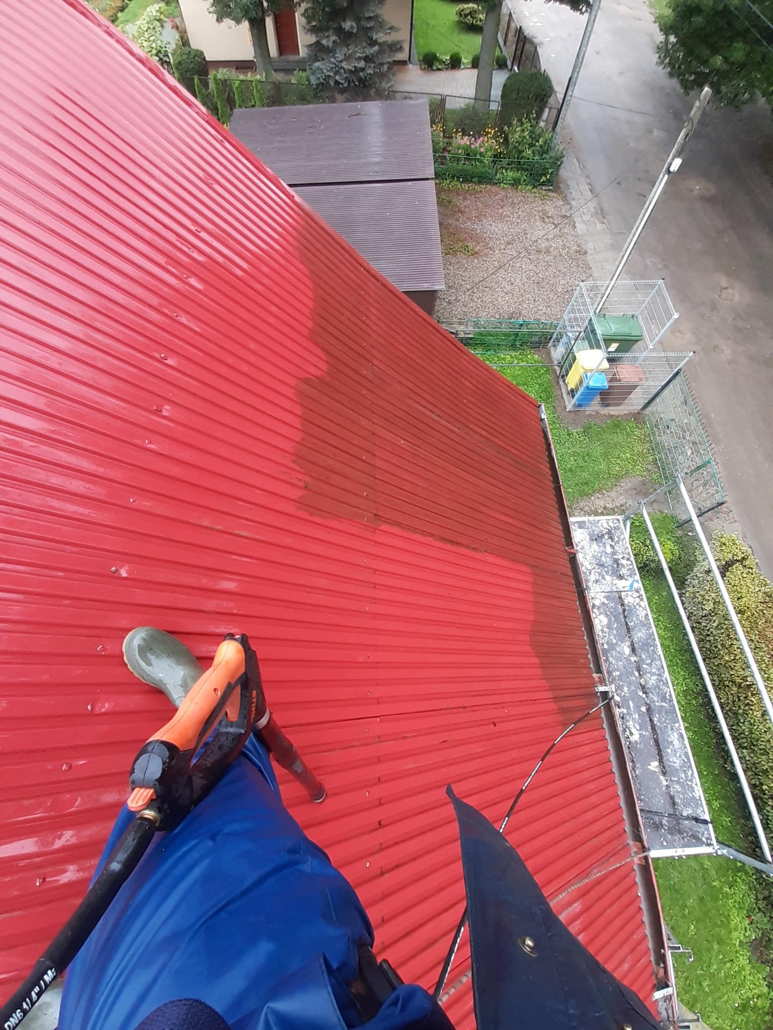 Czerwony, blaszany dach w trakcie czyszczenia myjką ciśnieniową, widoczne różnice w czystości powierzchni, osoba w niebieskim kombinezonie i gumowym bucie na dachu, z góry widok na ulicę i ogród
