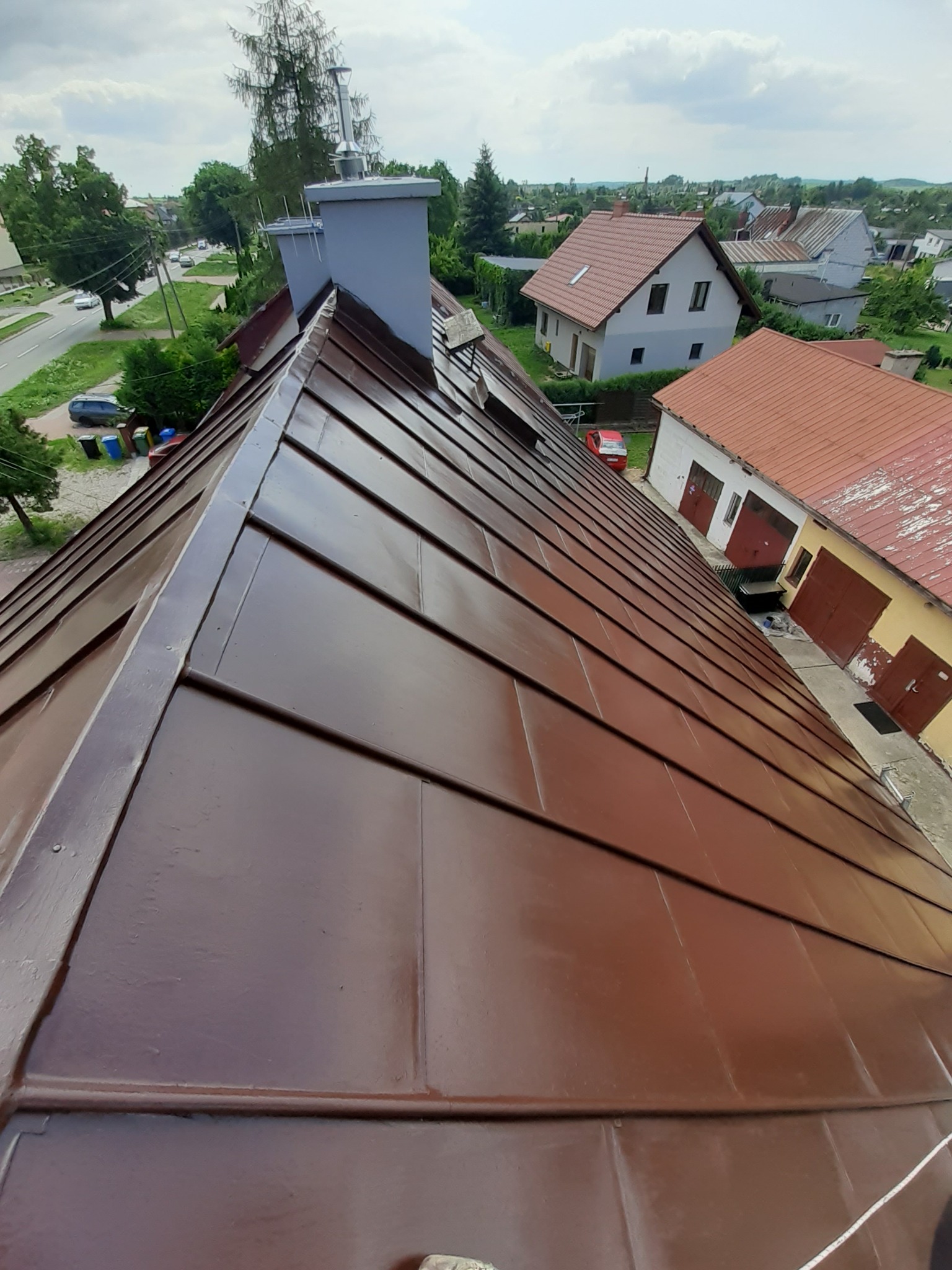 Widok z góry na brązowy, metalowy dach z kominem na tle zabudowań mieszkalnych i zieleni w słoneczny dzień.
