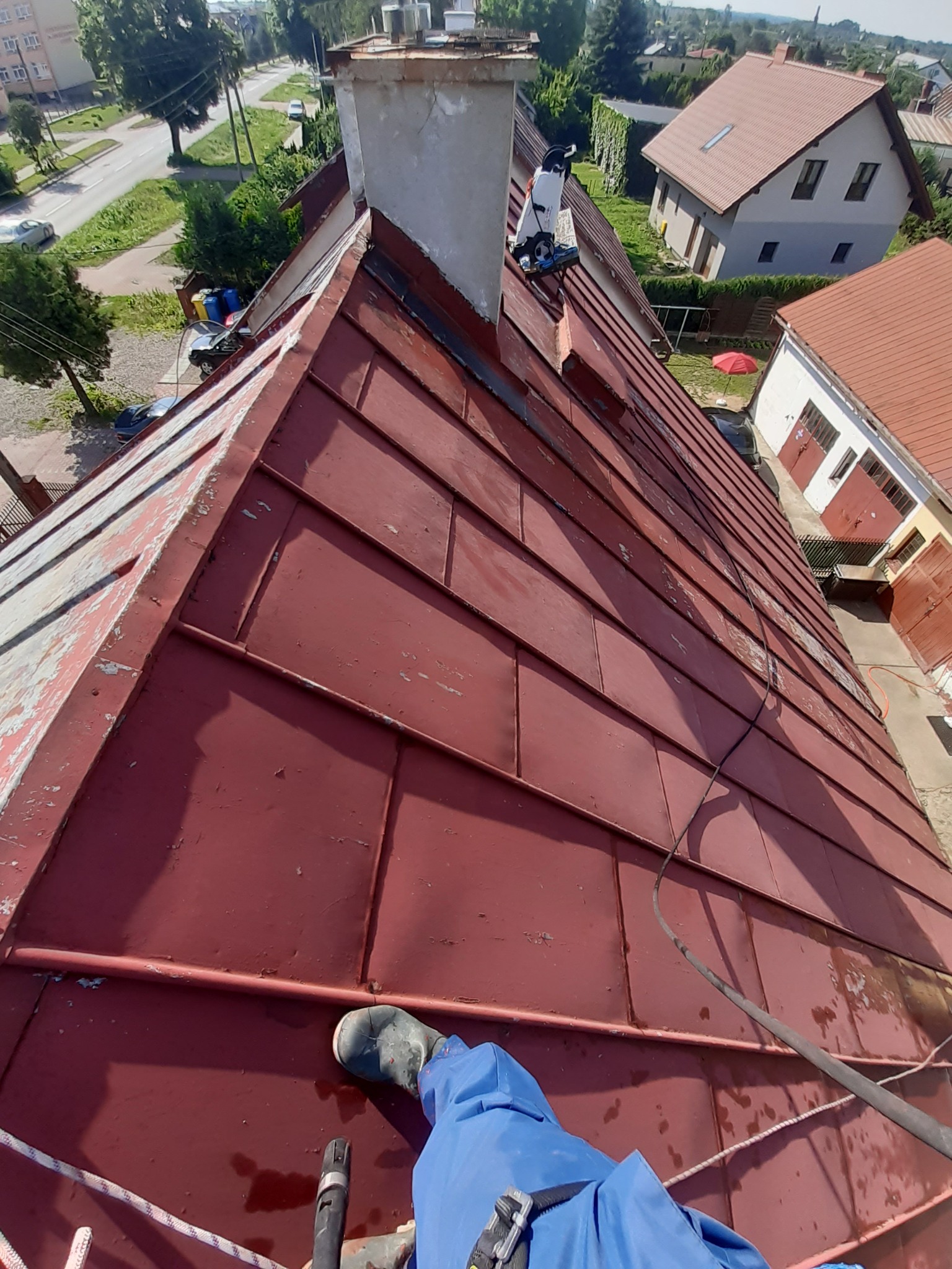 Czerwony, metalowy dach z widocznymi śladami korozji podczas mycia pod ciśnieniem, z fragmentem stroju roboczego i buta osoby wykonującej prace oraz sprzętem do mycia ciśnieniowego w tle.