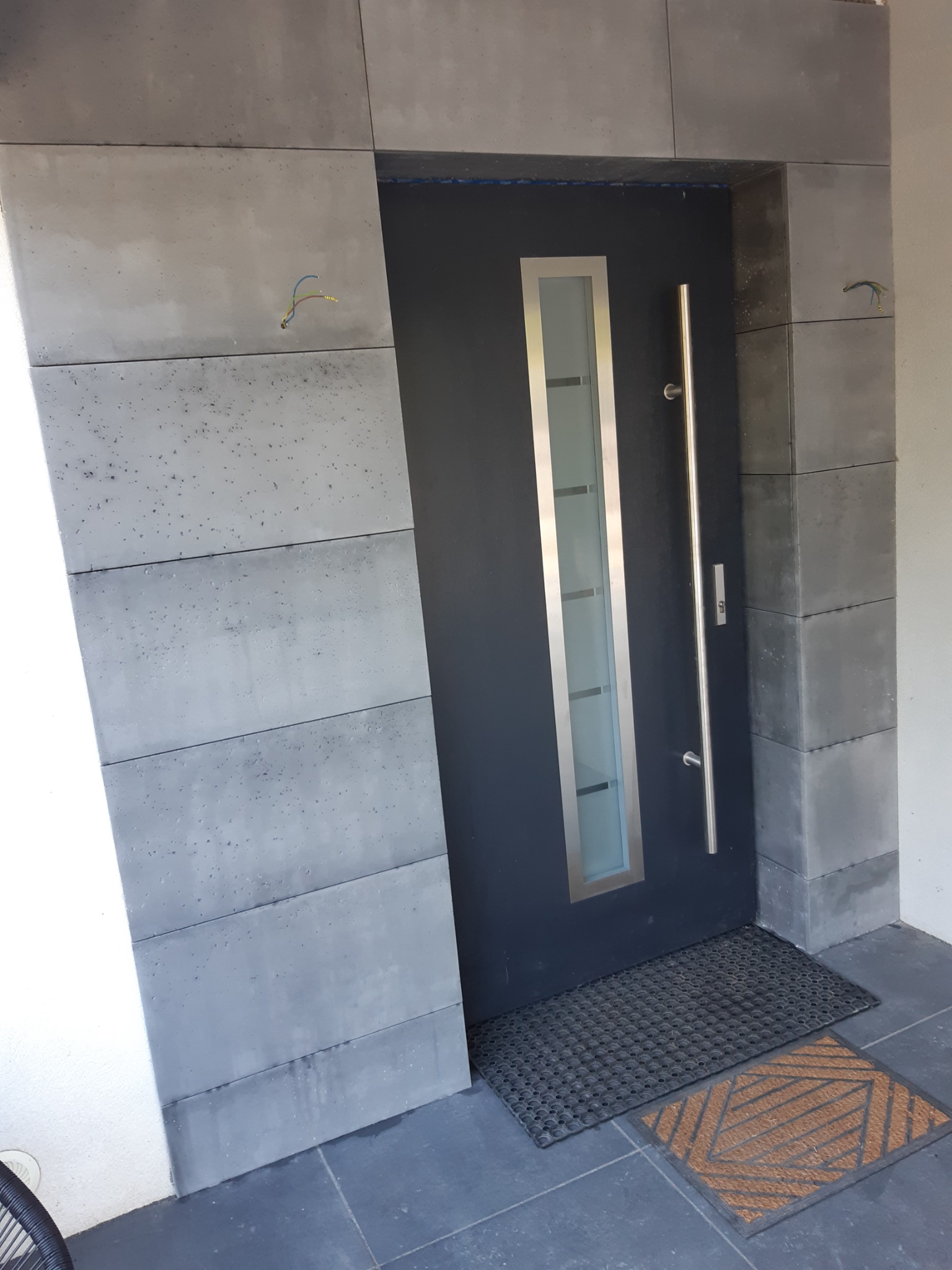 Nowoczesne wejście do domu z szarą, przeszkloną stolarką drzwiową i okładziną z betonowych płyt. Widoczne wystające kable elektryczne w ścianie.