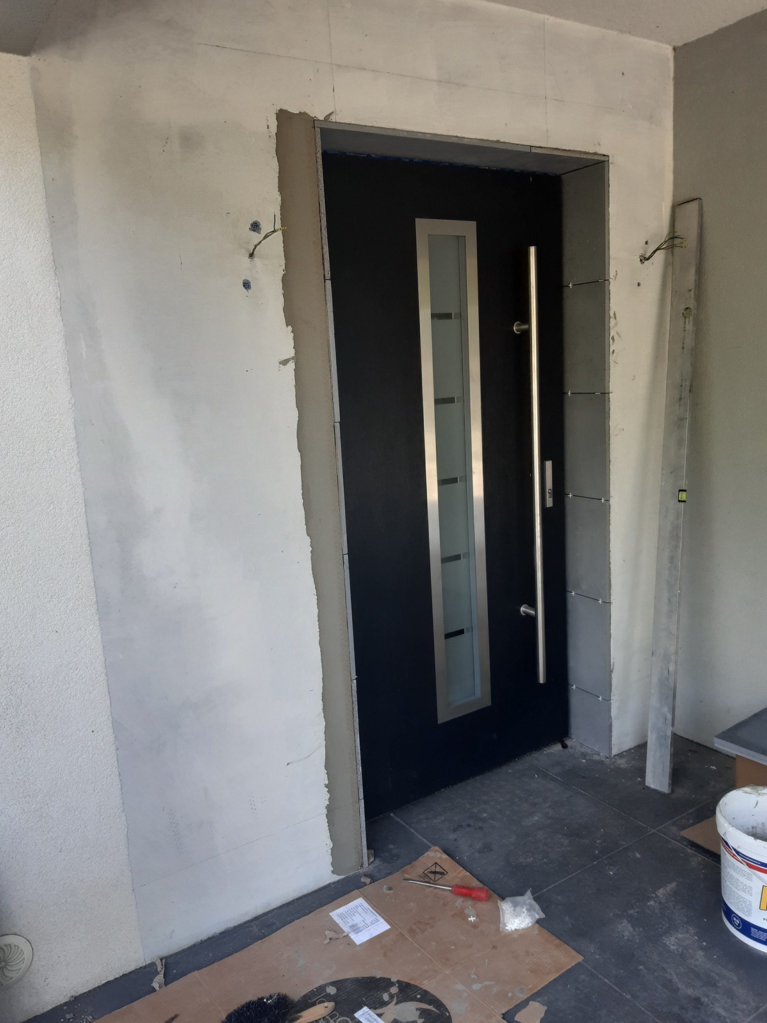 Montaż drzwi zewnętrznych: czarne drzwi z długim, srebrnym uchwytem, widoczne prace wykończeniowe wokół ościeżnicy, surowa ściana z wystającymi kablami, poziomica oparta o ścianę, narzędzia...