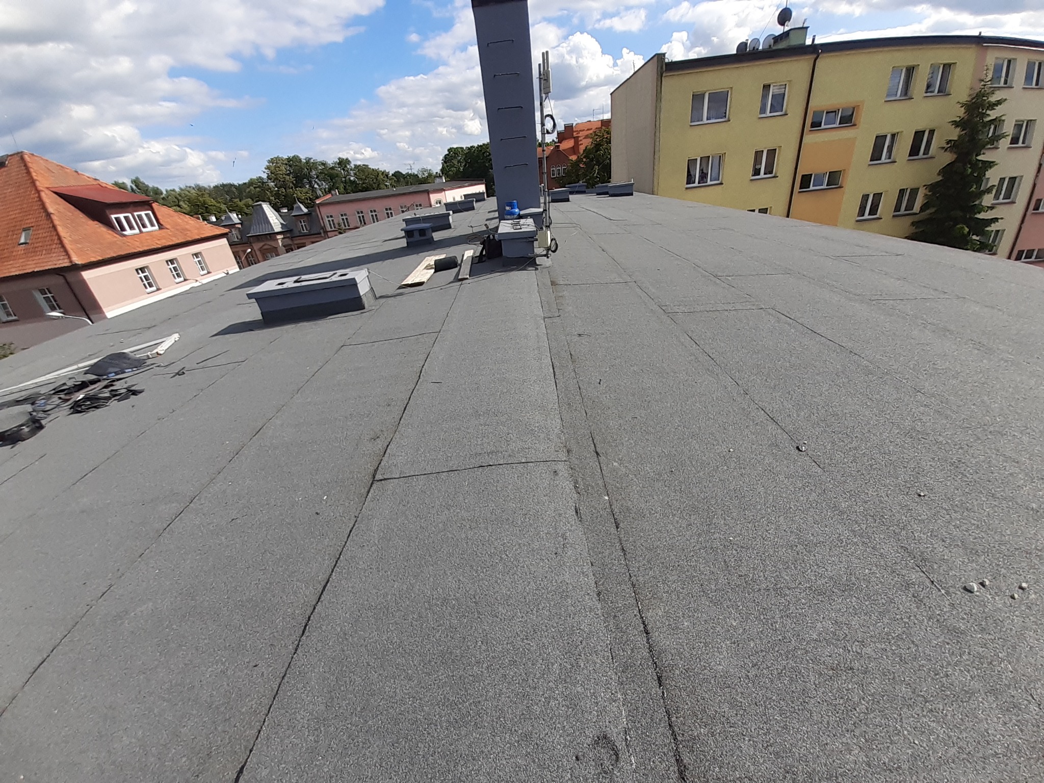Wykończony, płaski dach pokryty papą termozgrzewalną z widocznymi kominami wentylacyjnymi i antenami, na tle zabudowy miejskiej w słoneczny dzień.