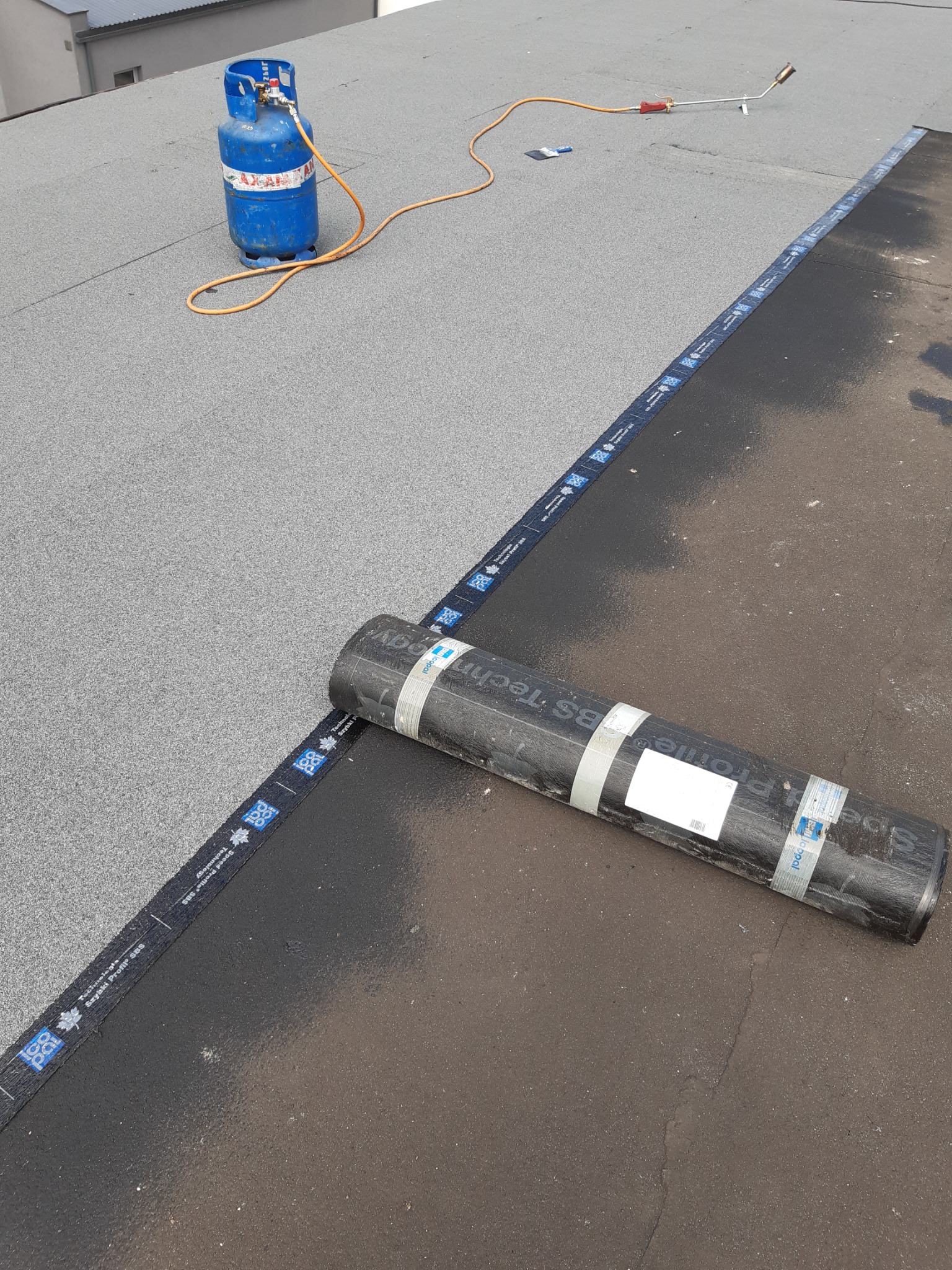 Prace dekarskie na dachu: rolka papy termozgrzewalnej, butla gazowa i palnik dekarski na płaskim dachu pokrytym szarą i czarną papą.