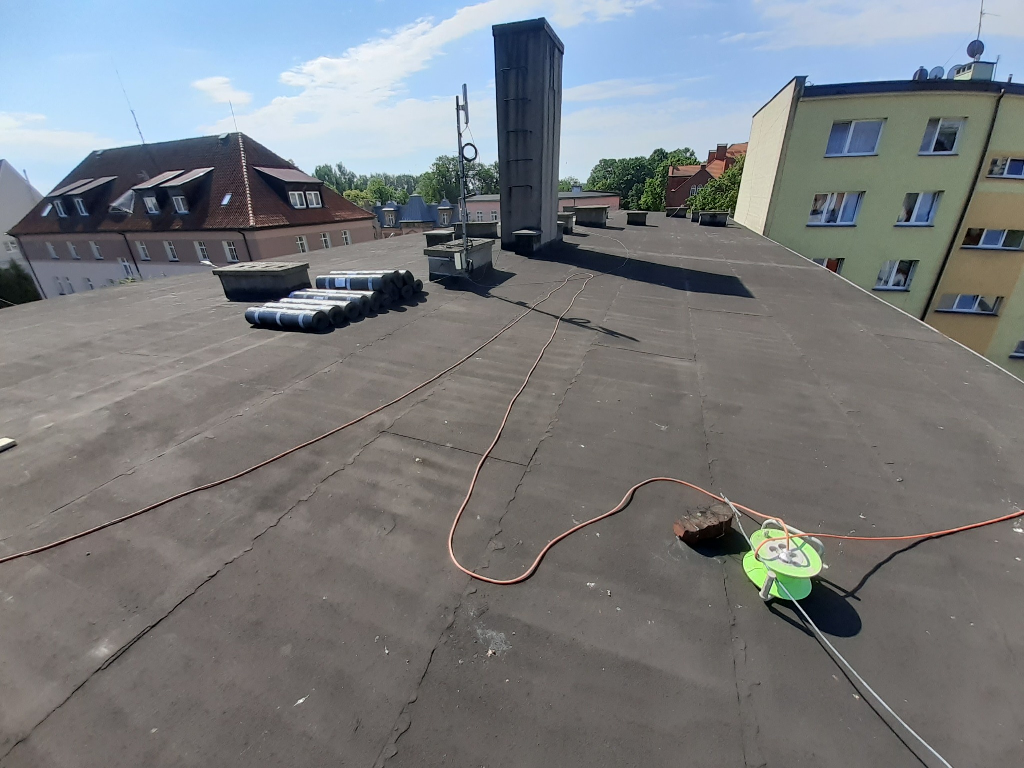 Widok z góry na płaski dach pokryty papą, z kominem, anteną, rolkami papy i zwiniętym kablem na zielonej szpuli, w tle budynki mieszkalne w słoneczny dzień.