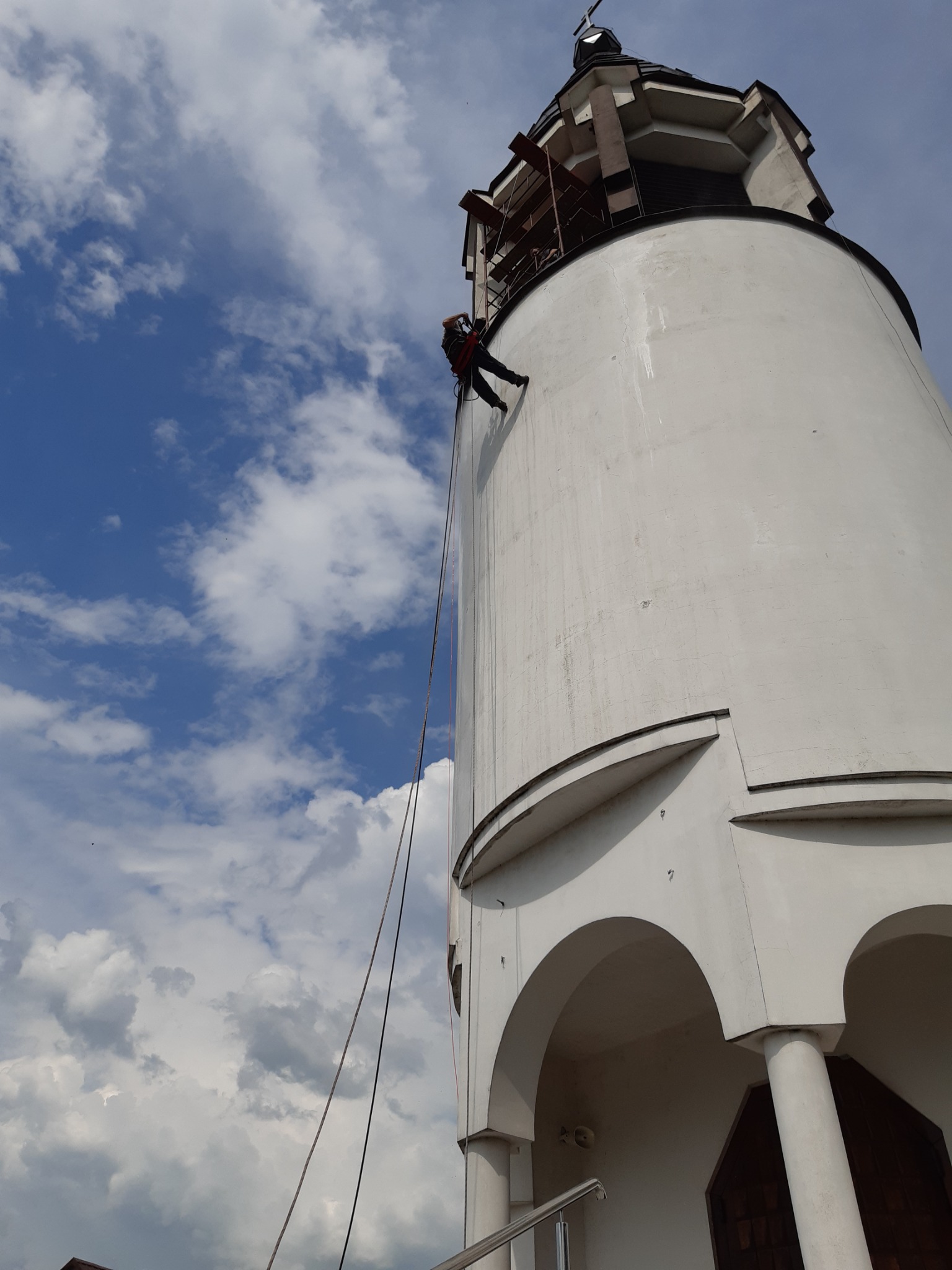 Pracownik na linie zabezpieczającej, w trakcie pracy na wysokościach, na tle białej wieży kościelnej i błękitnego nieba z chmurami.