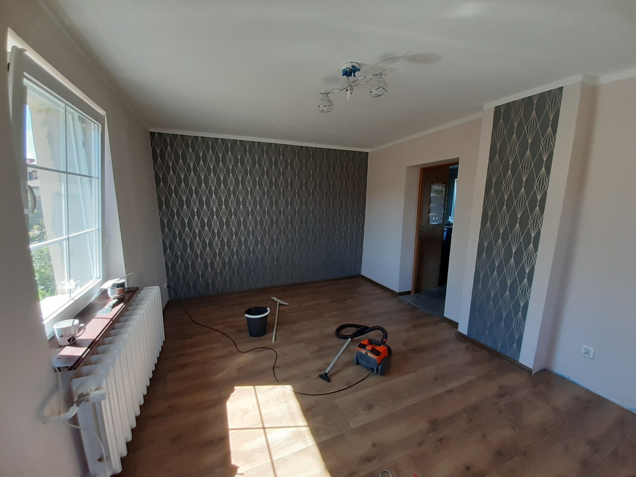 Pokój po remoncie z nową podłogą panelową, wzorzystą tapetą na ścianach, grzejnikiem pod oknem i sprzętem do sprzątania.