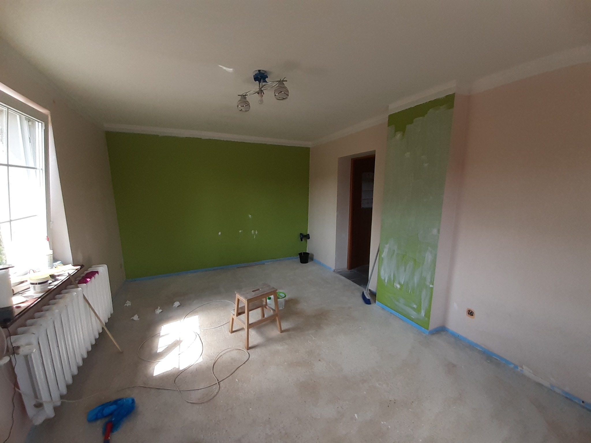 Pokój w trakcie malowania: jedna ściana zielona, druga różowa, widoczny stołek malarski, pędzel przy grzejniku i kabel na podłodze.