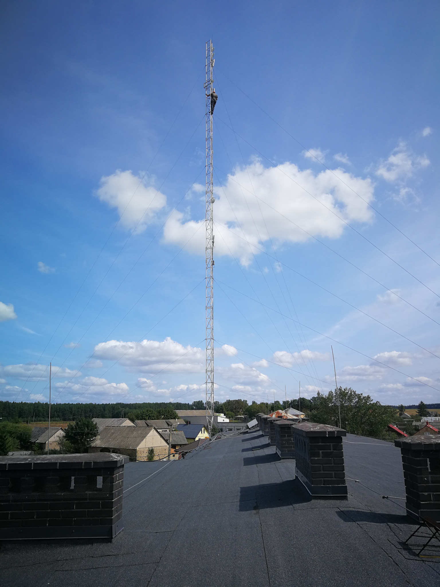 Praca na wysokościach w Olecku: Mężczyzna na wysokiej wieży telekomunikacyjnej widoczny z dachu budynku z kominami na tle błękitnego nieba z chmurami.