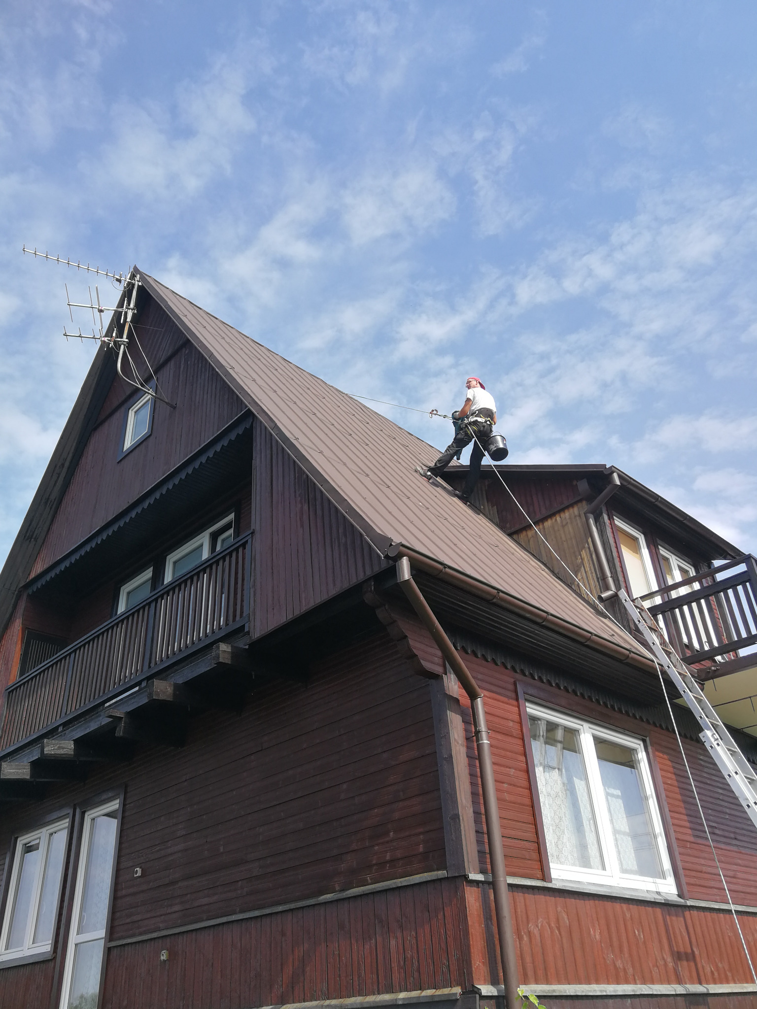 Pracownik w uprzęży na spadzistym dachu drewnianego domu z widocznymi rynnami, antena telewizyjna na szczycie, drabina oparta o ścianę, błękitne niebo w tle.