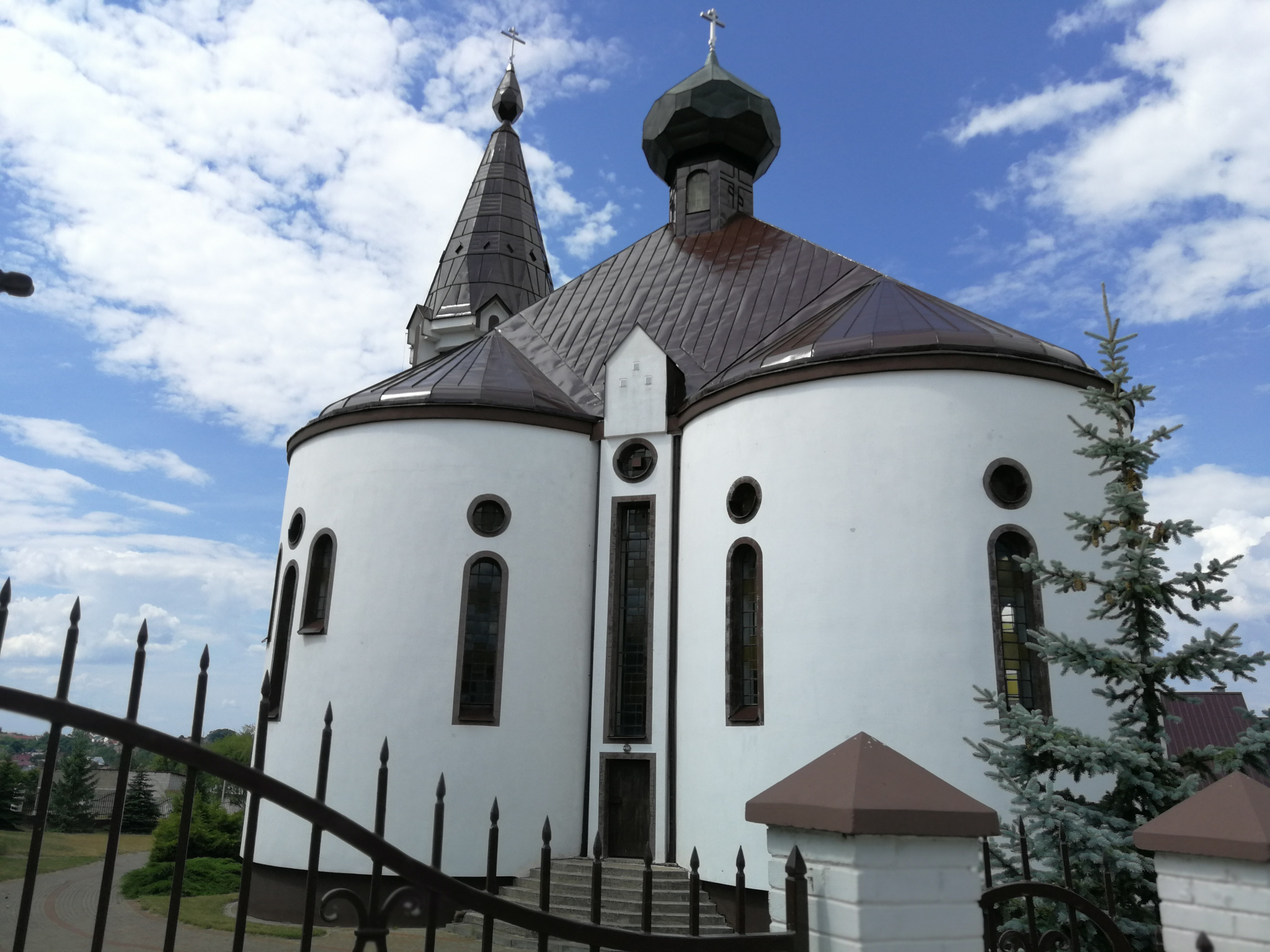 Biała cerkiew z brązowym dachem i dwiema wieżami zwieńczonymi krzyżami, widziana zza metalowego ogrodzenia, na tle błękitnego nieba z obłokami.