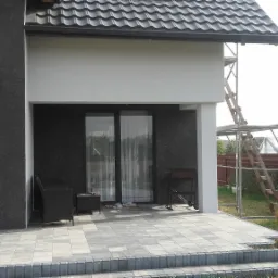 Nowoczesny dom z ciemną elewacją, jasnym podcieniem i szarą kostką brukową na tarasie, z widocznym rusztowaniem i fragmentem huśtawki ogrodowej.