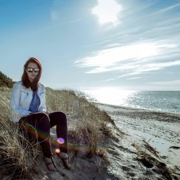 MCF STUDIO - Kobieta w okularach przeciwsłonecznych siedzi na wydmie, ubrana w białą kurtkę i ciemne spodnie, z widokiem na morze w słoneczny dzień.