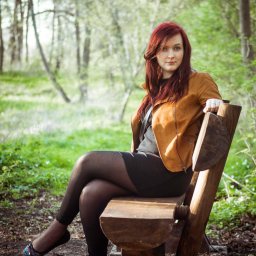 MCF STUDIO - Kobieta z rudymi włosami siedzi na drewnianej ławce w parku, ubrana w czarną sukienkę, rajstopy i brązową kurtkę, z nogami skrzyżowanymi w kostkach, w tle widoczne drzewa i zieleń.