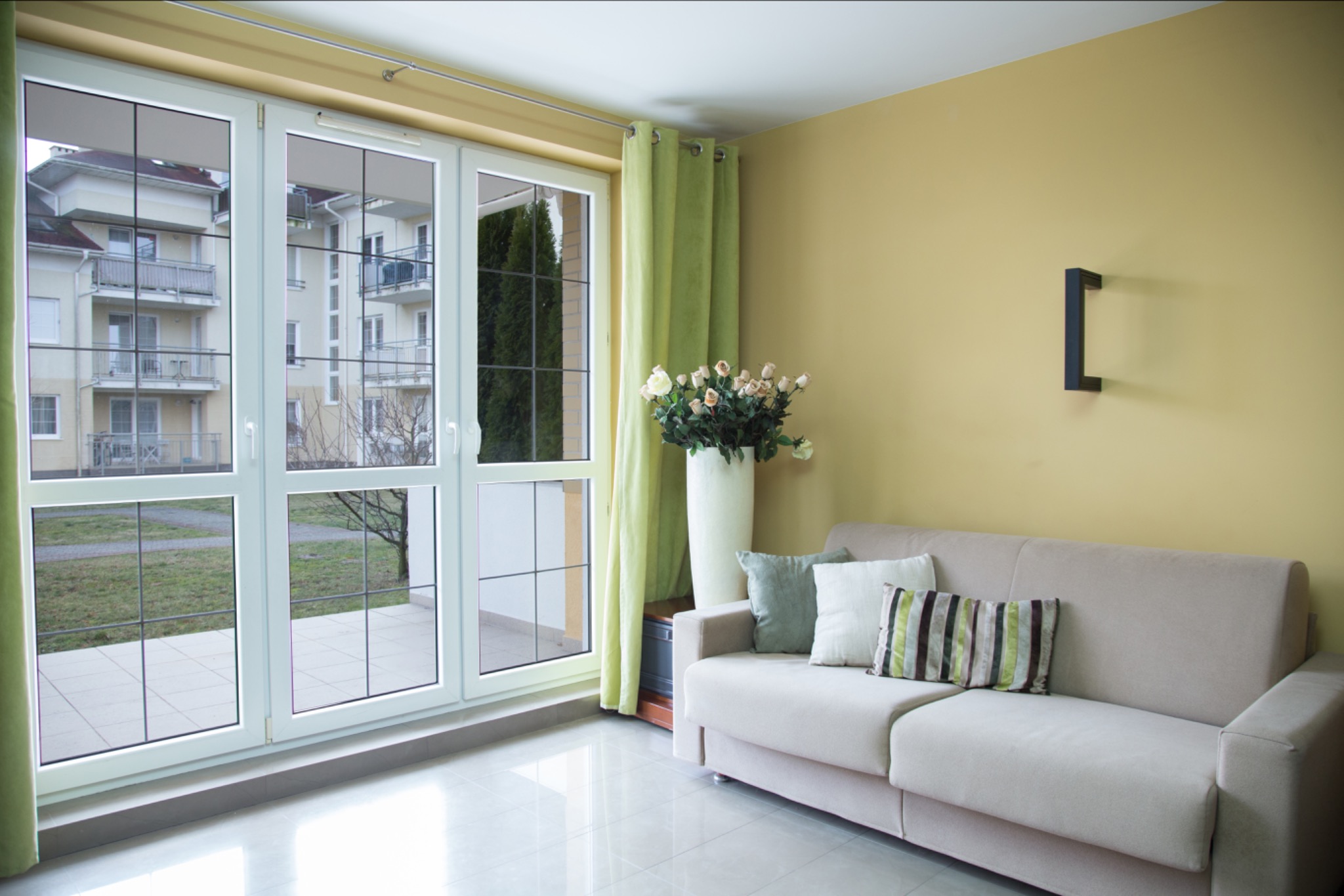 Jasne wnętrze pokoju z dużym oknem, beżową sofą z poduszkami, wazonem z kwiatami i zielonymi zasłonami, widok na budynek z balkonami przez okno.