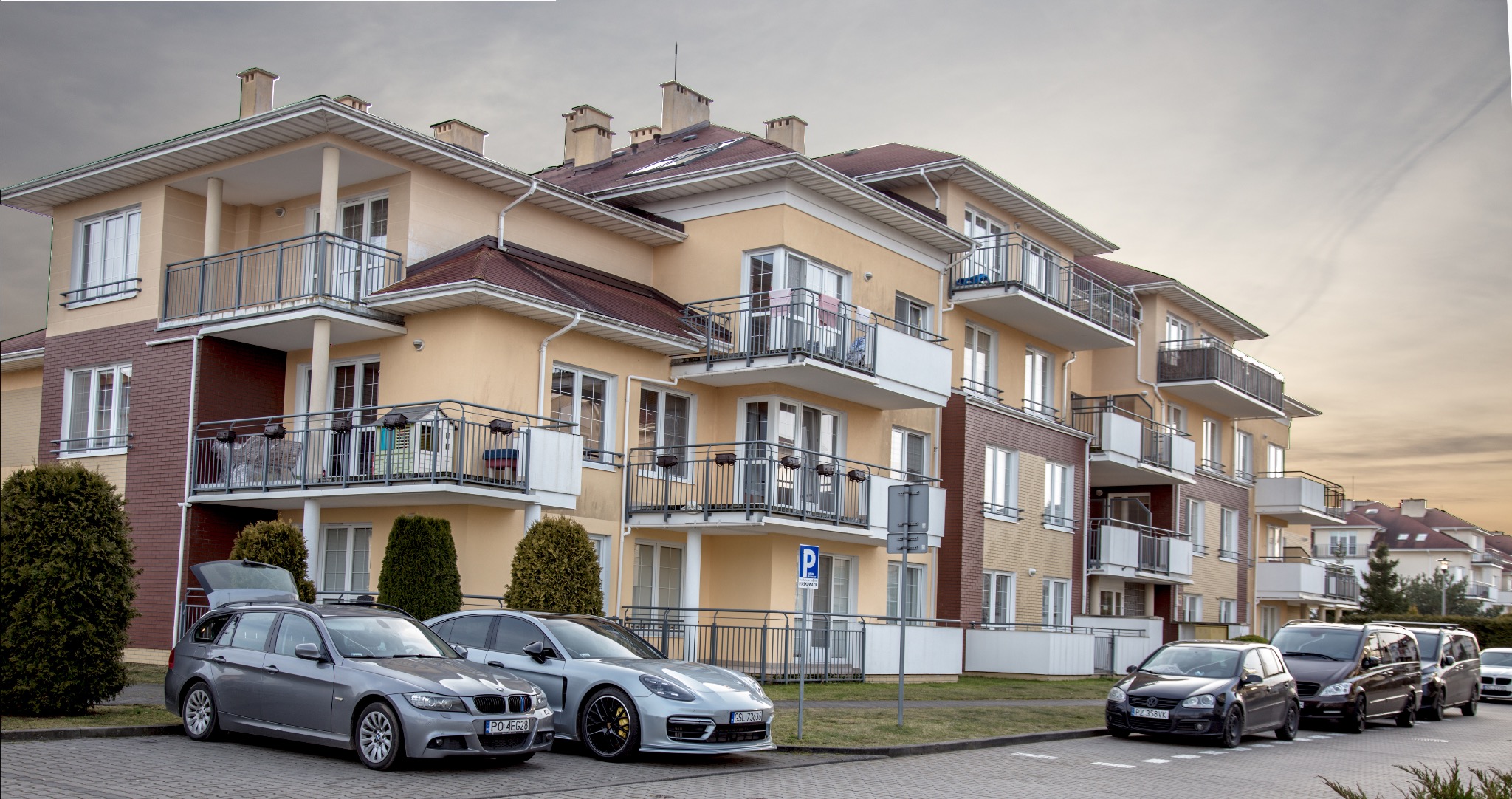 Nowoczesny budynek mieszkalny z balkonami i zaparkowanymi samochodami wzdłuż ulicy, widoczny znak parkingu.