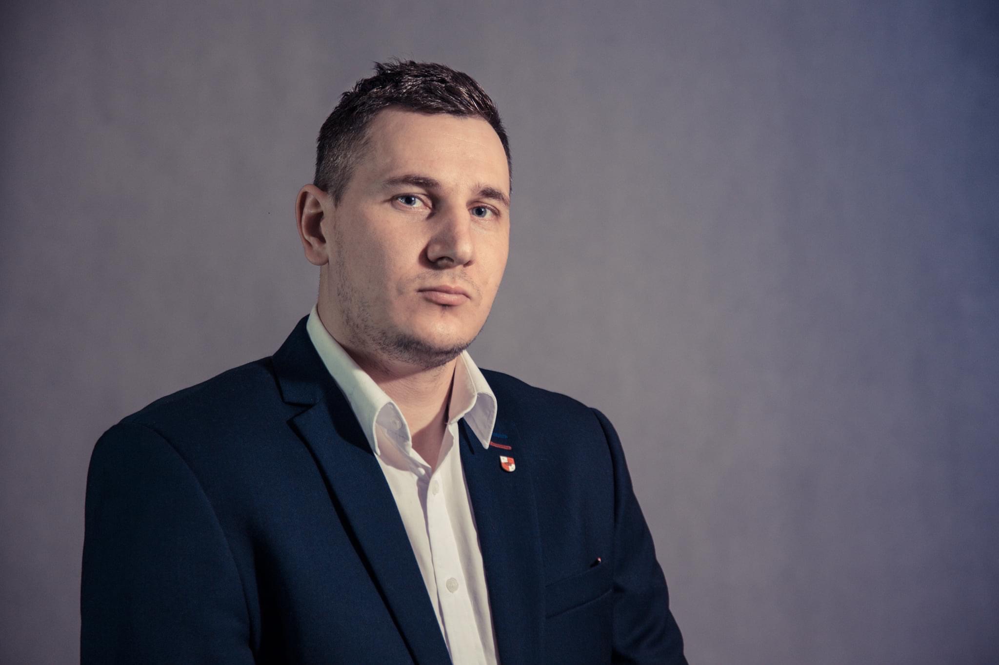 Portret mężczyzny w ciemnej marynarce i białej koszuli, z przypinką na klapie, na szarym tle.