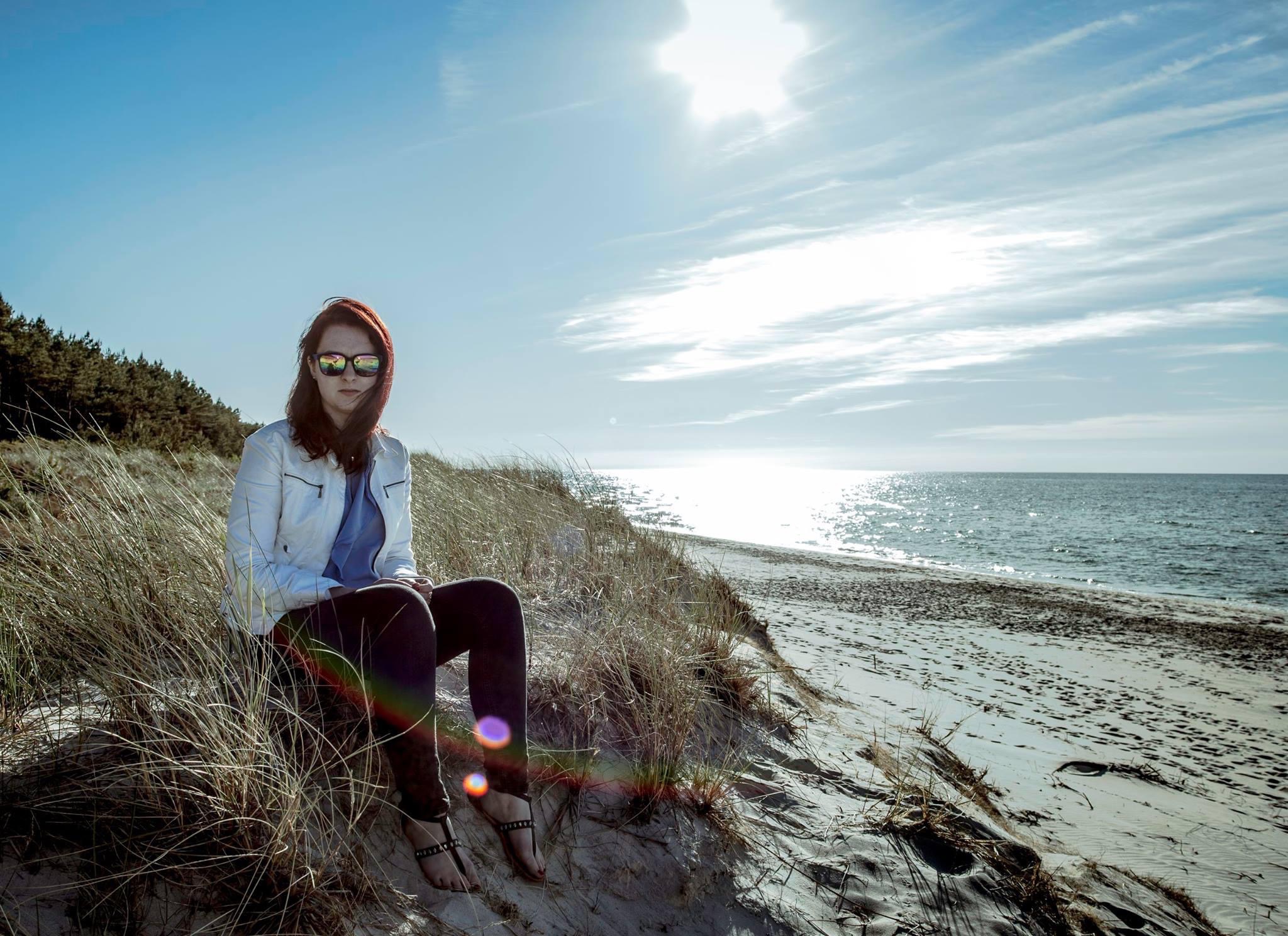 Kobieta w okularach przeciwsłonecznych siedzi na wydmie, ubrana w białą kurtkę i ciemne spodnie, z widokiem na morze w słoneczny dzień.