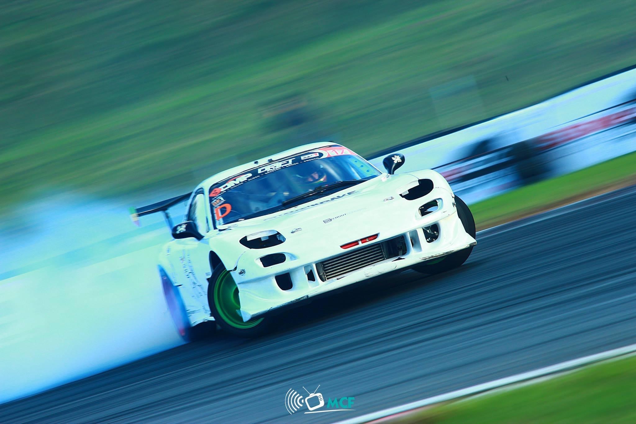 Biały samochód sportowy w poślizgu na torze wyścigowym, z zielonymi felgami i dymem unoszącym się z opon, uchwycony w dynamicznym ujęciu z efektem rozmycia ruchu.