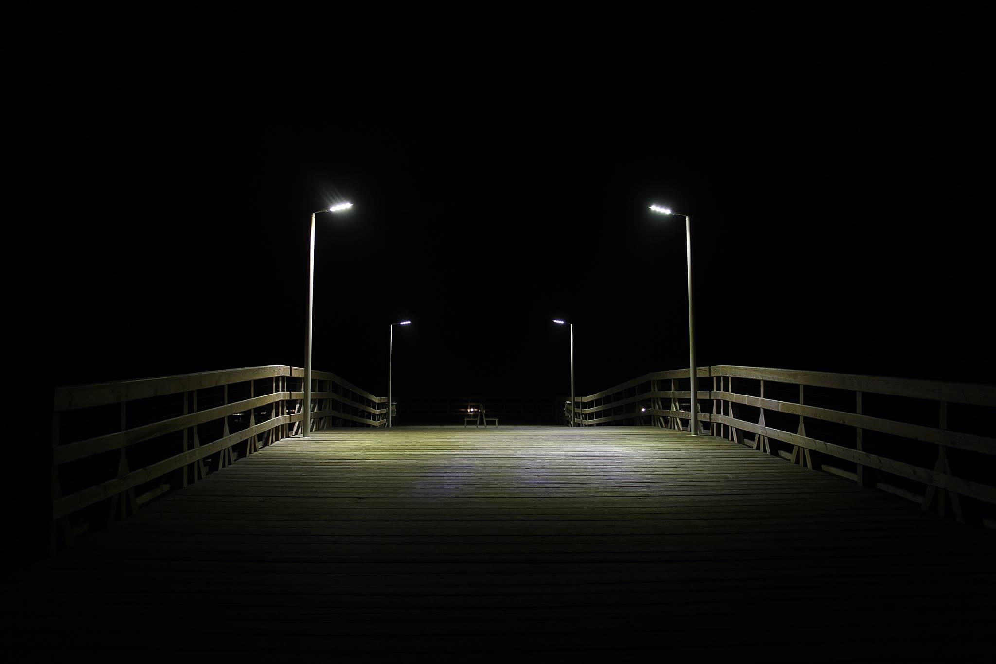 Drewniany pomost oświetlony lampami ulicznymi w nocnej scenerii, prowadzący w kierunku dwóch krzeseł w oddali.