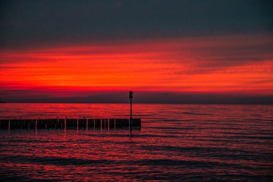 Intensywny czerwony zachód słońca nad spokojnym morzem, z drewnianym pomostem wystającym z wody, na którym siedzą ptaki.