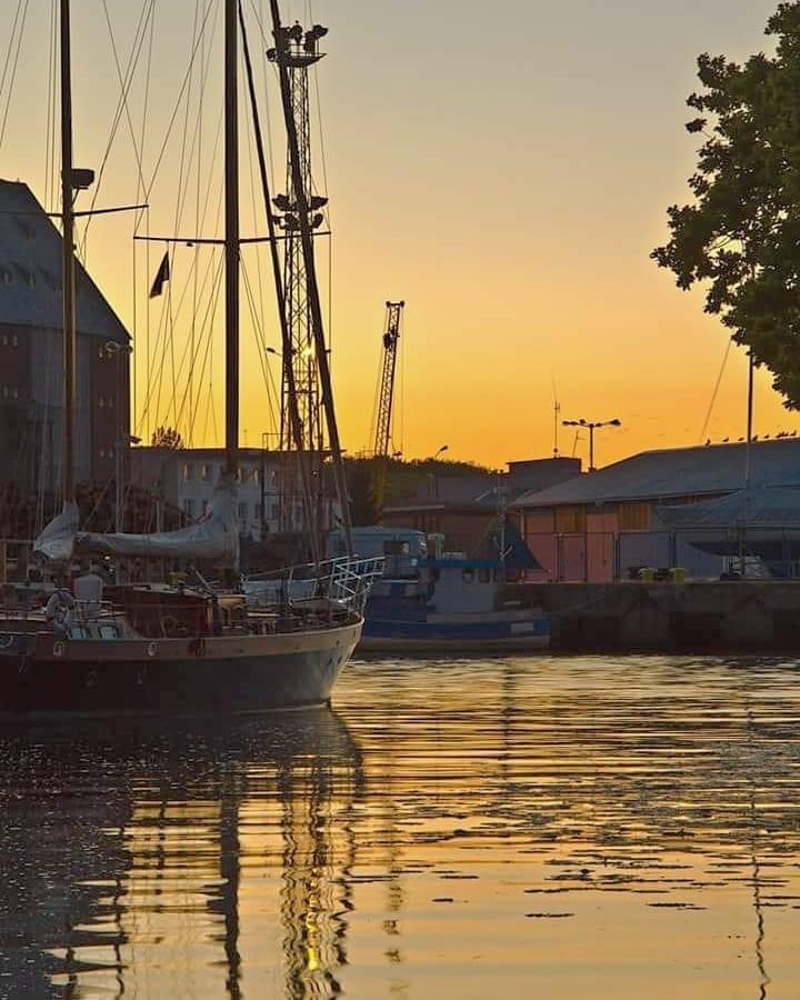 Żaglówka przy nabrzeżu o zmierzchu, odbicie w wodzie, w tle budynki portowe i dźwigi.