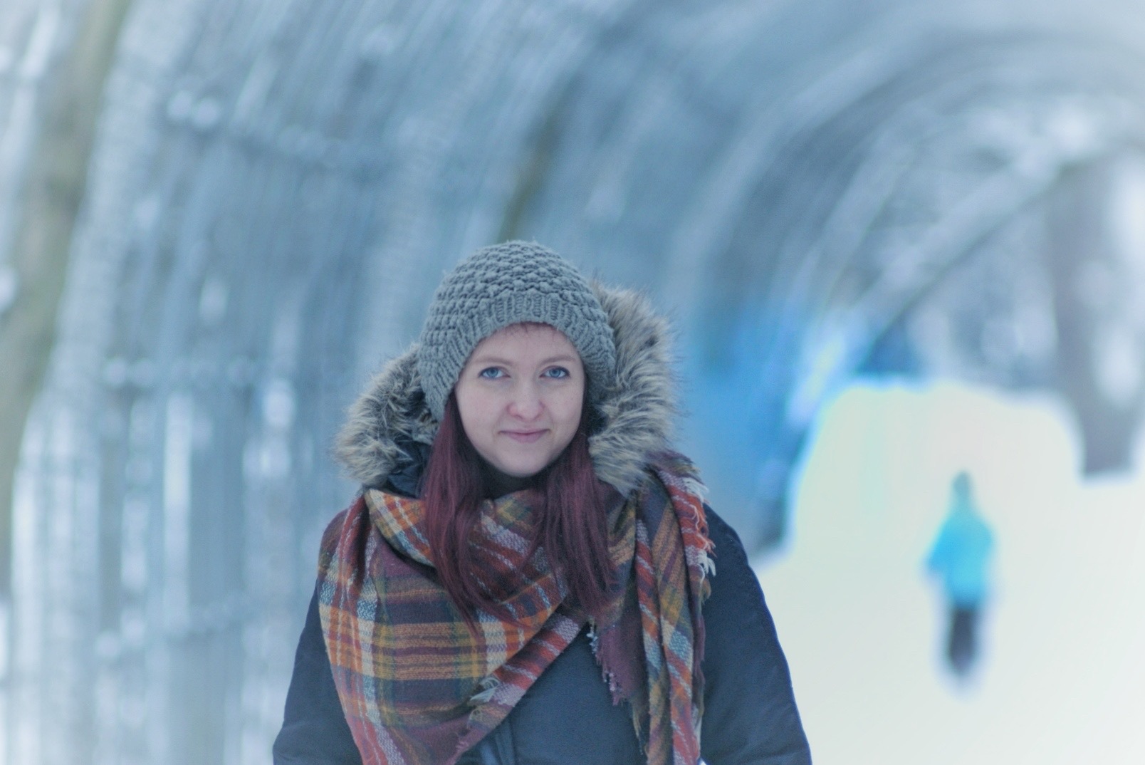 Portret kobiety w zimowej scenerii, ubrana w szarą czapkę z futrzanym pomponem i duży, kraciasty szal, na rozmytym tle tunelu z lodowych konstrukcji.
