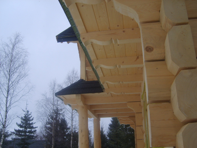 Fragment konstrukcji domu z bali z widocznymi drewnianymi belkami, filarami i elementami dachu, na tle zimowego krajobrazu z drzewami bez liści.