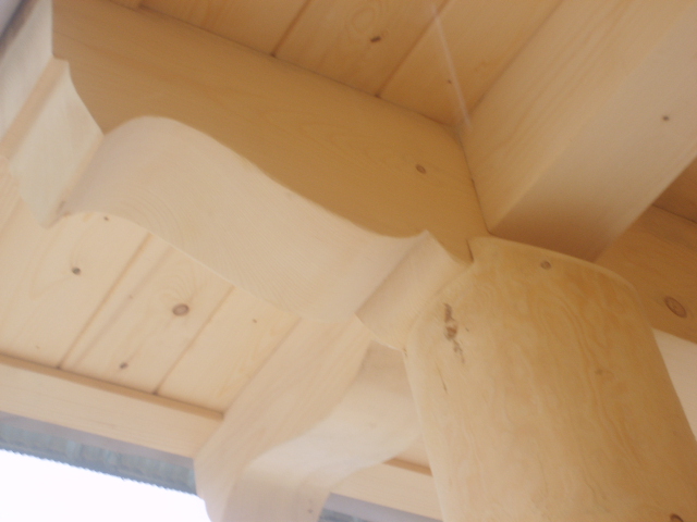 Fragment konstrukcji drewnianej z okrągłym słupem i dekoracyjnymi wycięciami, widok od dołu na jasne drewno.