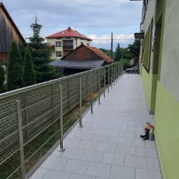 Długi balkon z szarymi płytkami i metalową konstrukcją zabezpieczającą, widok na okoliczne domy i zieleń, puszka farby i karton na podłodze.