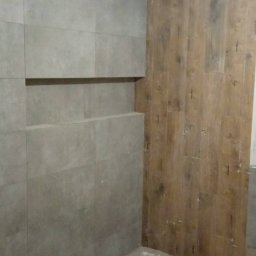 Łukasz Leman - Ściana łazienki wyłożona szarymi płytkami z wnęką, sąsiadująca z fragmentem ściany wykończonej panelami imitującymi drewno. Podłoga zabezpieczona kartonem.