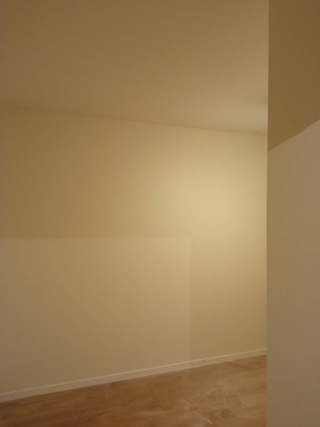 Pomalowane na beżowo ściany i sufit w pustym pokoju z drewnianą podłogą, widoczna listwa przypodłogowa.