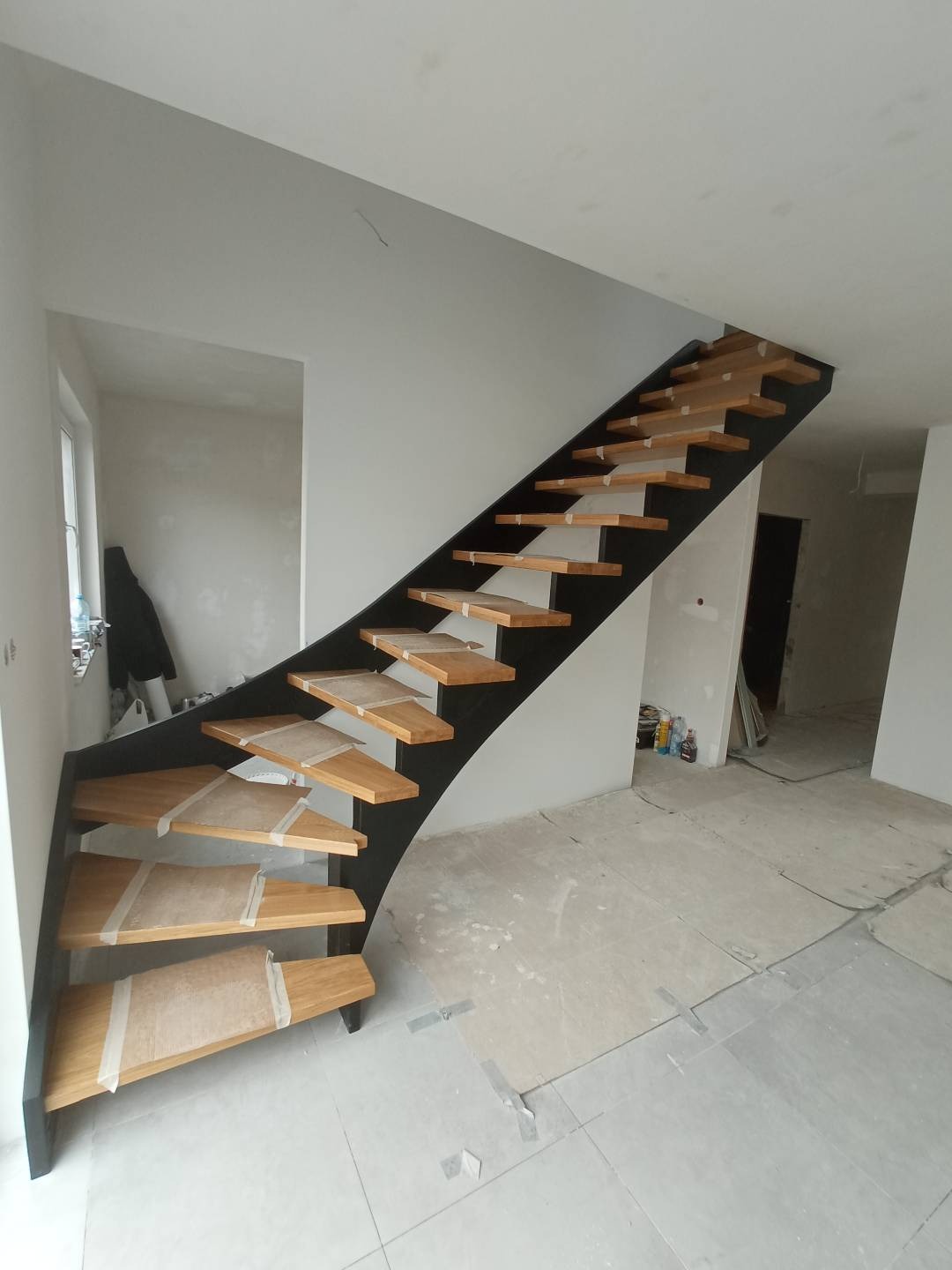 Nowoczesne schody drewniane z czarną metalową konstrukcją, zabezpieczone folią ochronną podczas prac wykończeniowych w jasnym wnętrzu.