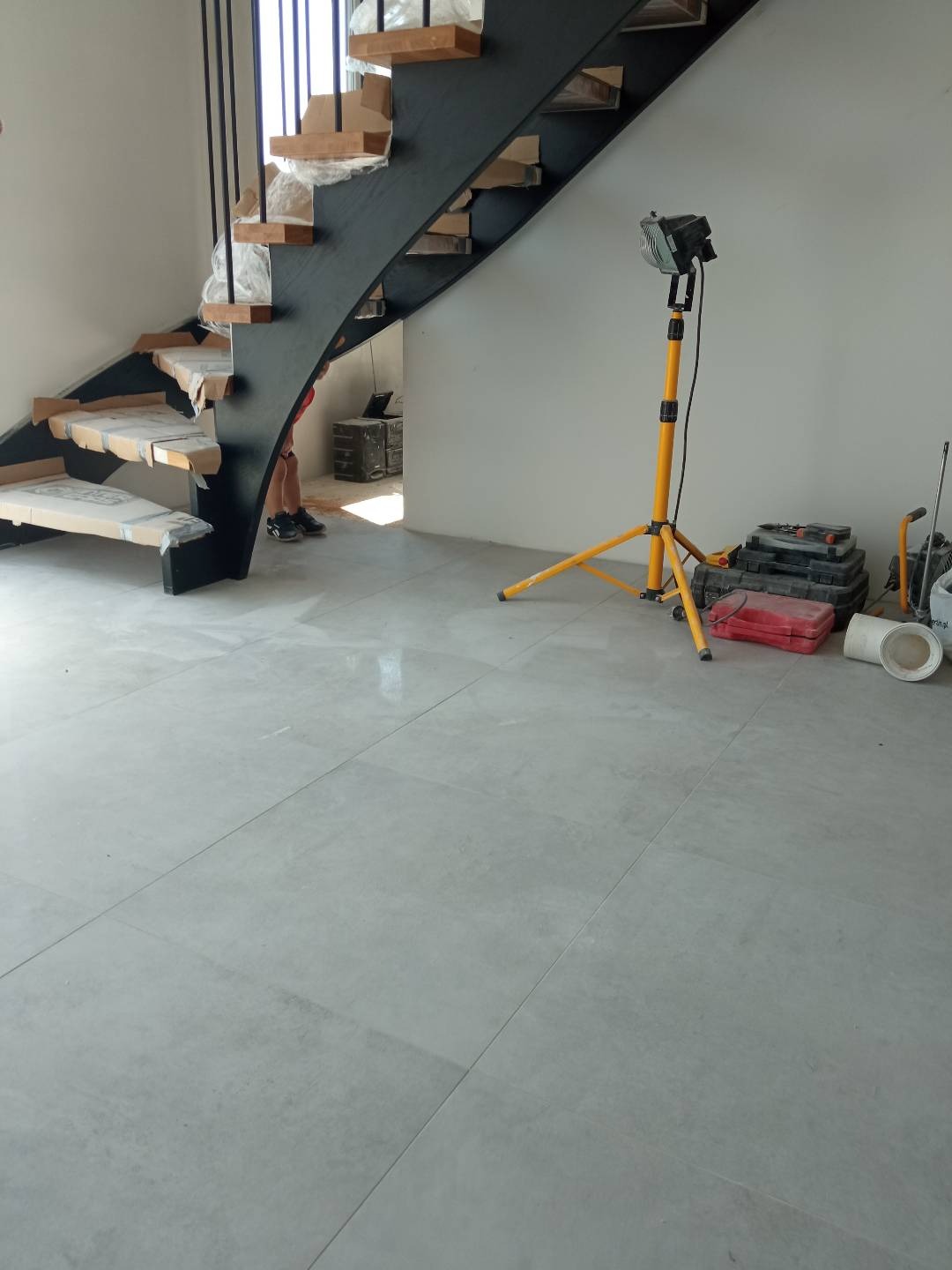 Montaż nowoczesnych schodów z drewnianymi stopniami i czarną metalową konstrukcją, zabezpieczonych kartonem, w jasnym wnętrzu z szarymi płytkami podłogowymi, oświetlonym lampą budowlaną.