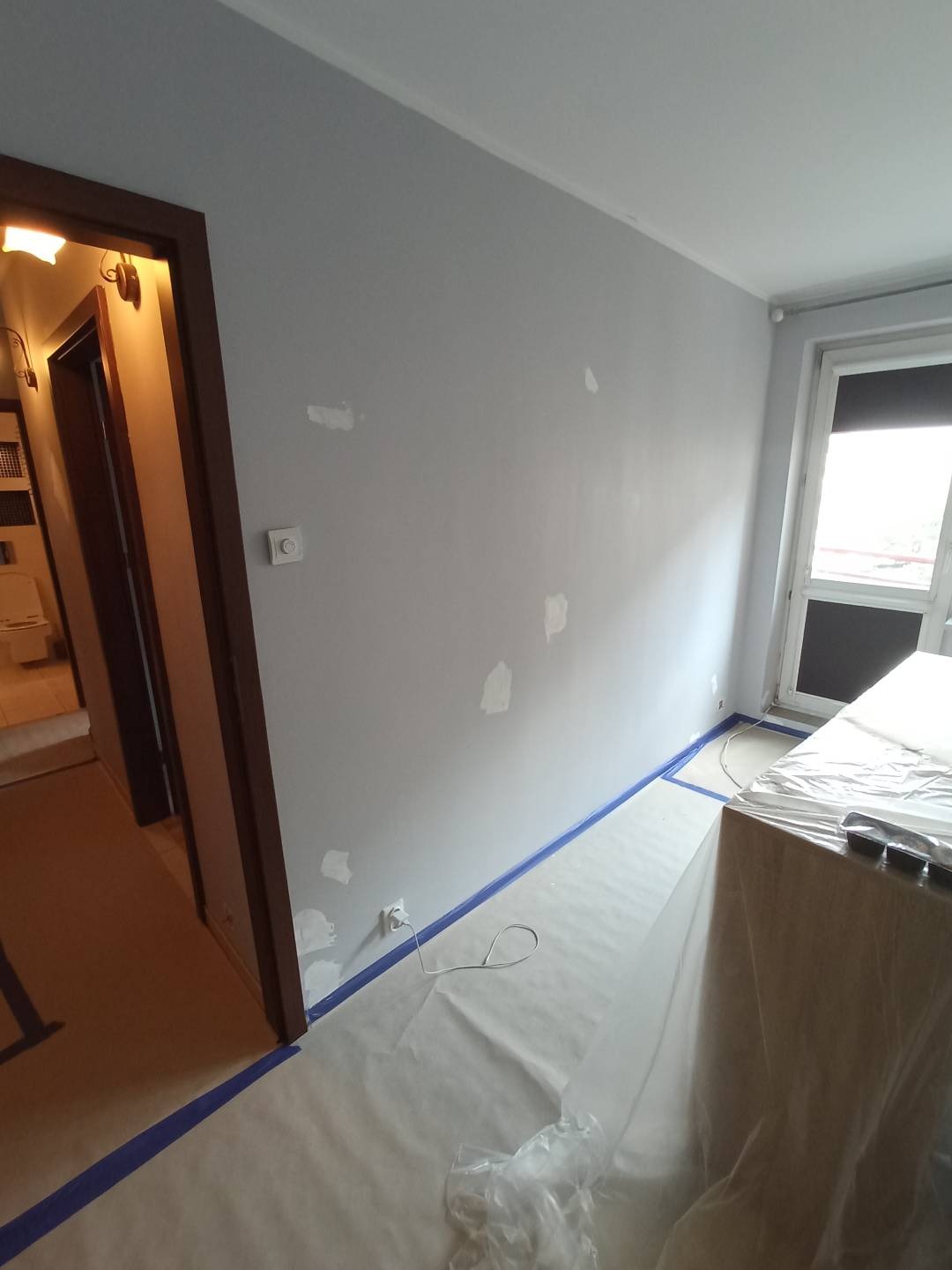 Przygotowanie ściany do malowania: szpachlowana, zagruntowana ściana w szarym odcieniu z widocznymi łatami szpachli, podłoga zabezpieczona folią i taśmą malarską, widoczny fragment okna i drzwi...