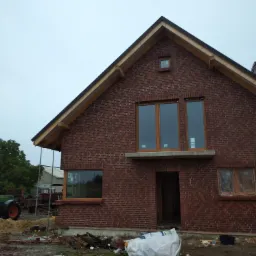 Surowy stan domu z czerwonej cegły z drewnianą konstrukcją dachu, widoczne okna z drewnianą ramą i niezabezpieczony otw&oacute;r drzwiowy, teren wok&oacute;ł z gruzem budowlanym i workiem na odpady.