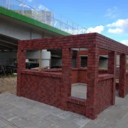 Niedokończona konstrukcja z czerwonej cegły, prawdopodobnie altana, na tle chodnika z kostki brukowej i wiaduktu z błękitnym niebem w tle.