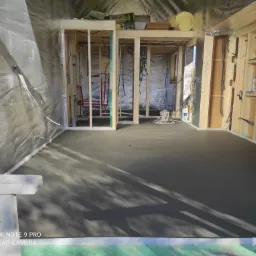 Świeżo wylana, gładka, szara posadzka w surowym wnętrzu budynku w trakcie budowy, widoczne drewniane szkielety ścian, izolacja folią aluminiową oraz instalacje.