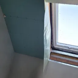 Montaż zielonej płyty gipsowo-kartonowej na suficie poddasza, widoczny otwór na instalację, okno dachowe zabezpieczone folią, ściany w trakcie przygotowania do malowania.