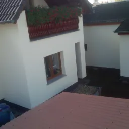 Biała elewacja domu z balkonem zdobionym czerwonymi kwiatami, widok z góry na brązowy dach i otoczenie.