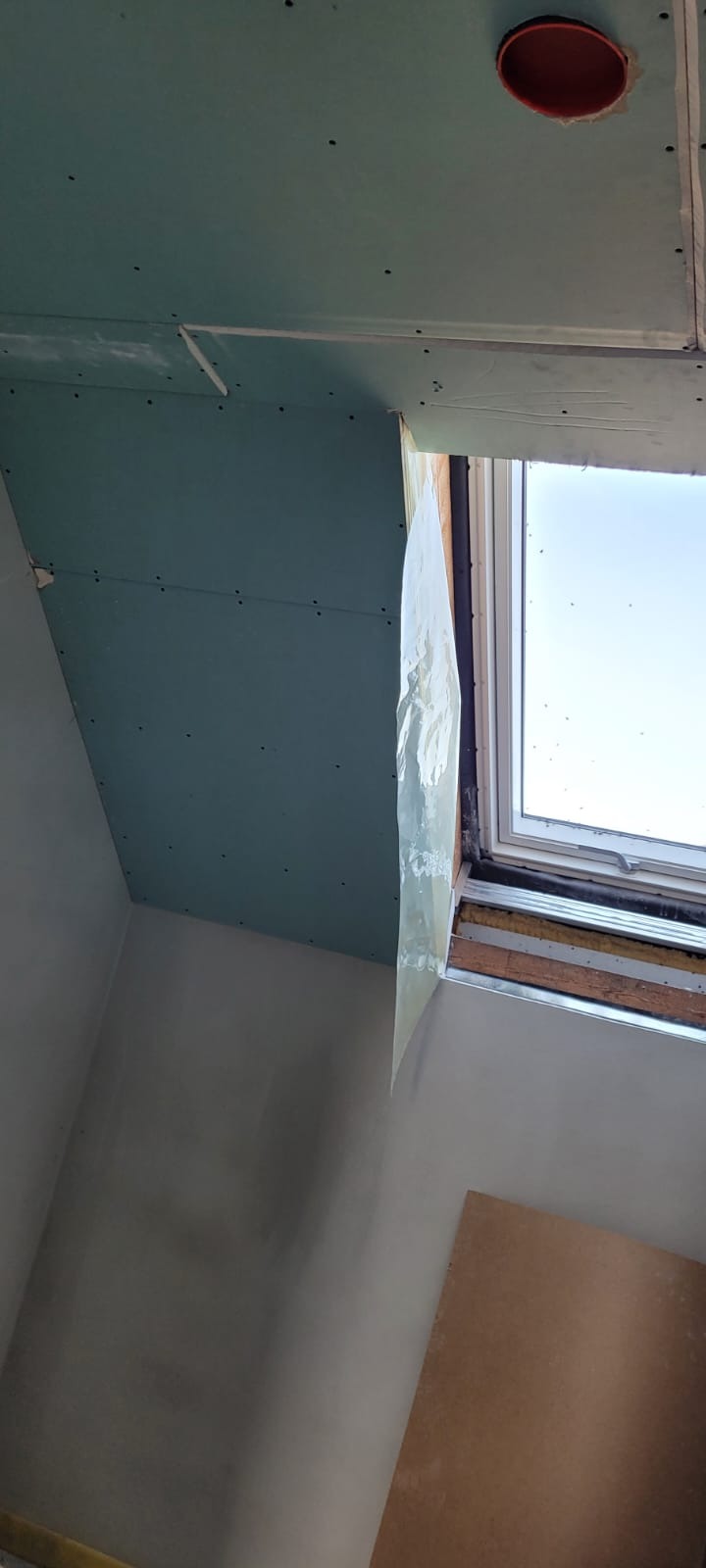 Montaż zielonej płyty gipsowo-kartonowej na suficie poddasza, widoczny otwór na instalację, okno dachowe zabezpieczone folią, ściany w trakcie przygotowania do malowania.