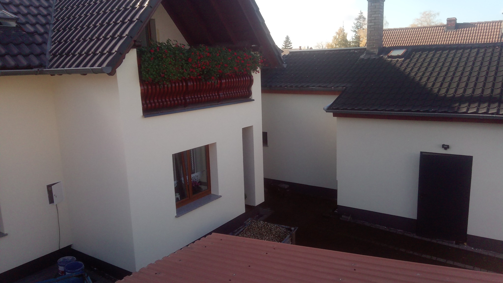 Widok z góry na budynek z jasną elewacją, drewnianym balkonem z czerwonymi kwiatami, ciemnym dachem i widocznymi elementami wykończeniowymi oraz pozostałościami po pracach malarskich.