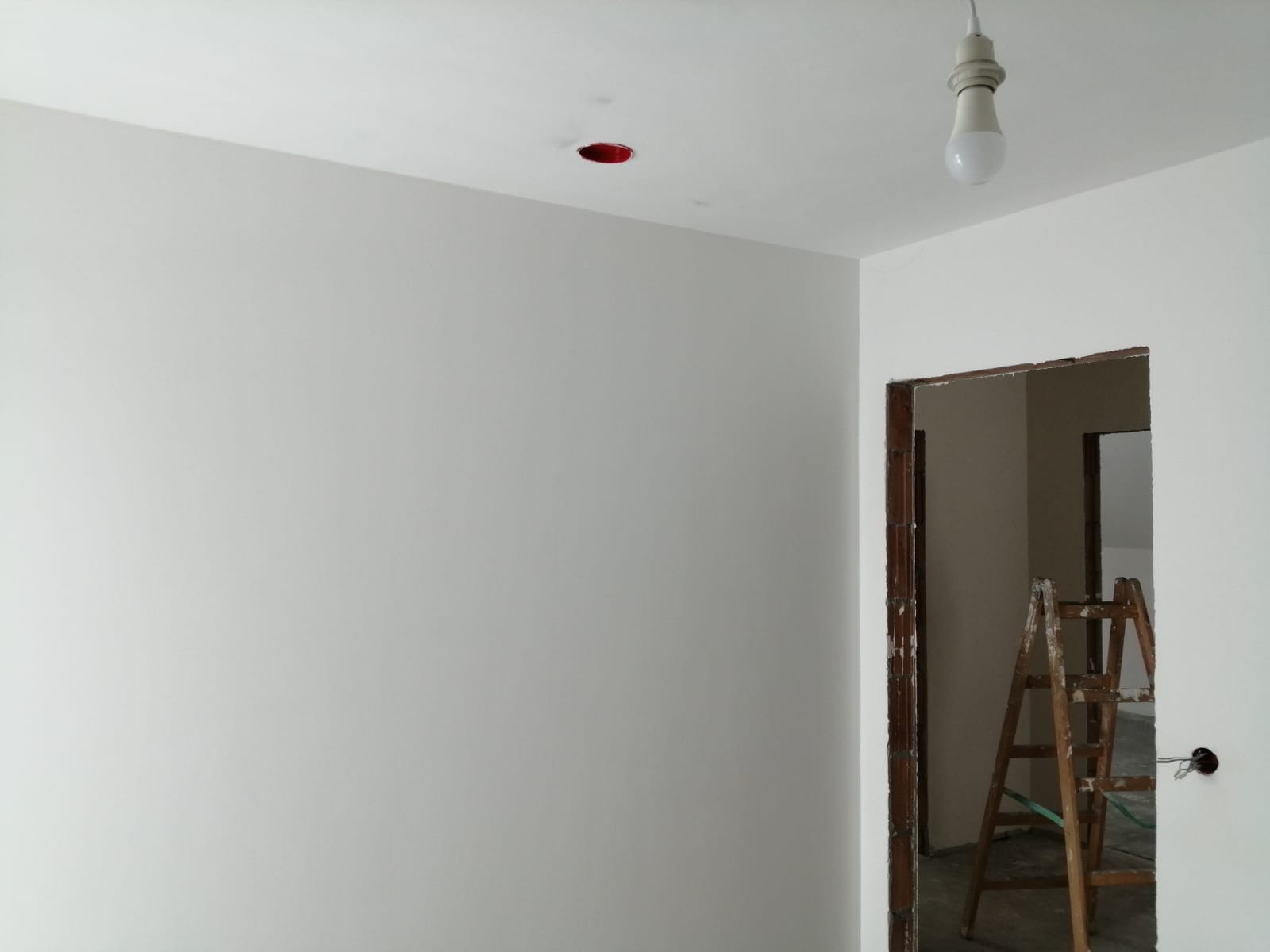 Świeżo pomalowane na biało wnętrze pokoju z widoczną drewnianą drabiną w przejściu, surową oprawą drzwi, otworem na lampę w suficie i wiszącą żarówką.