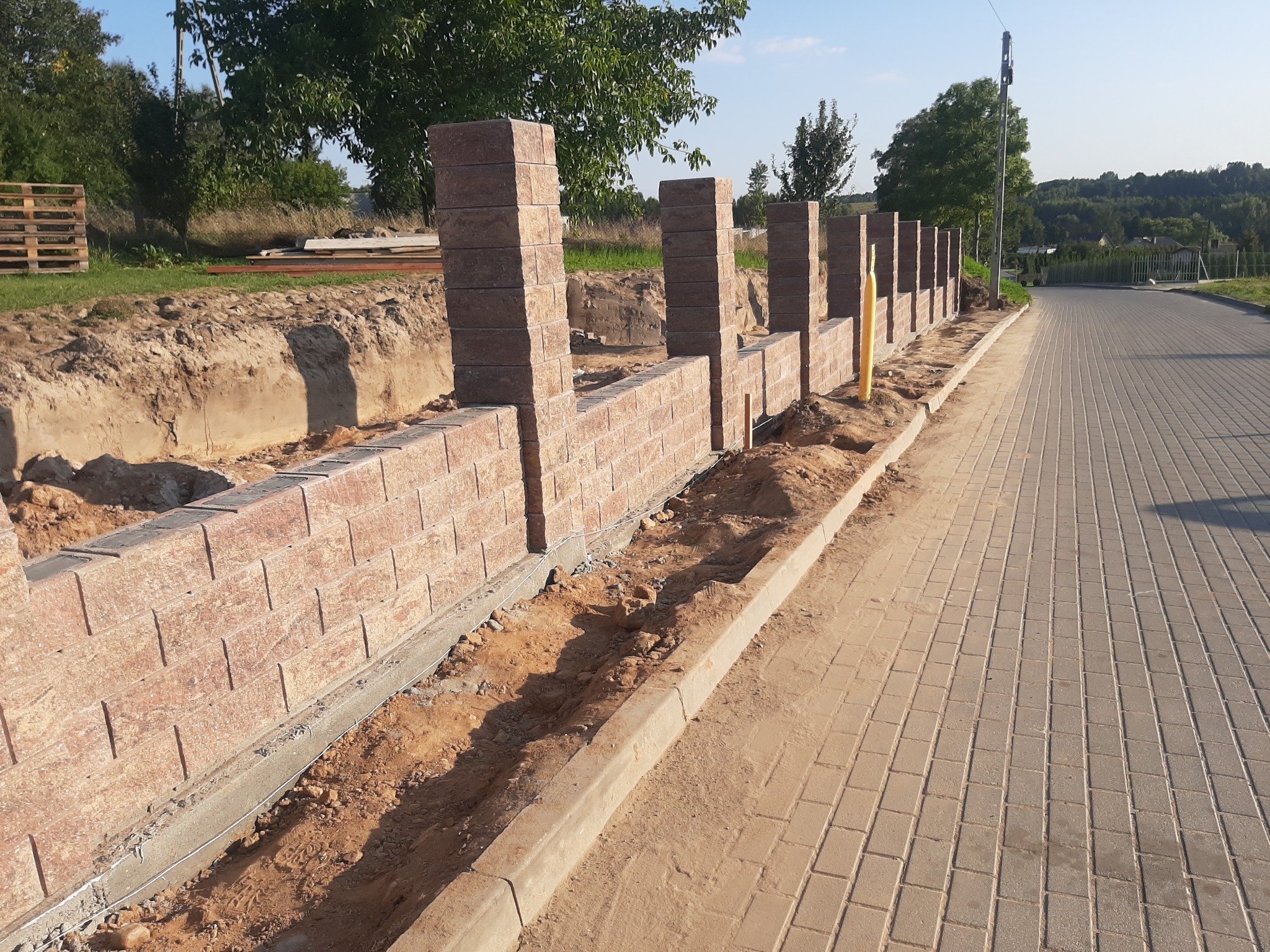 Wznoszenie ogrodzenia modułowego z cegieł klinkierowych, widoczne słupki i fragmenty muru, obok ułożona kostka brukowa, w tle zieleń.