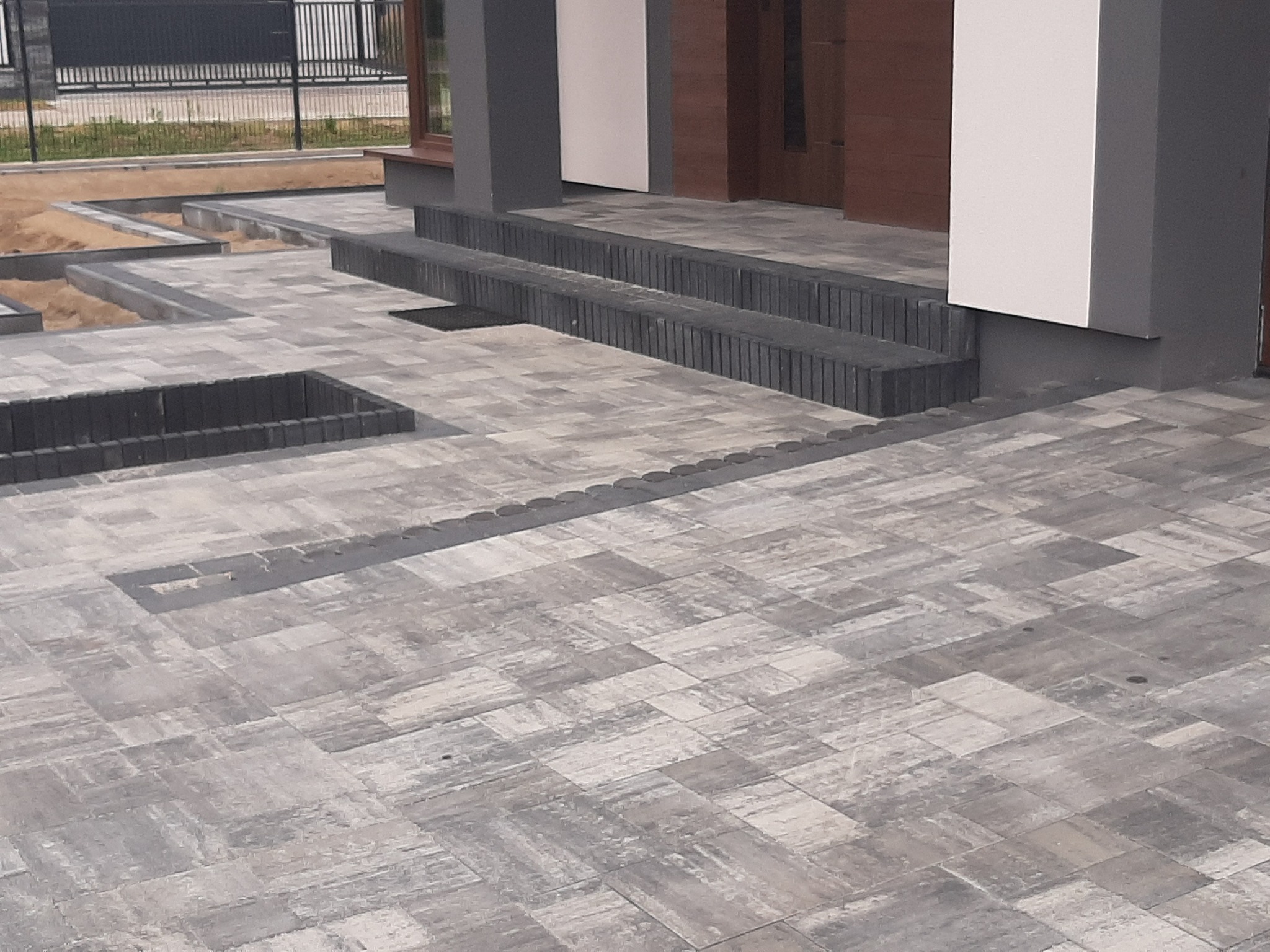 Ułożona szara kostka brukowa przed nowoczesnym domem, z niskimi schodami i rabatami z kostki, widoczny fragment ogrodzenia w tle.