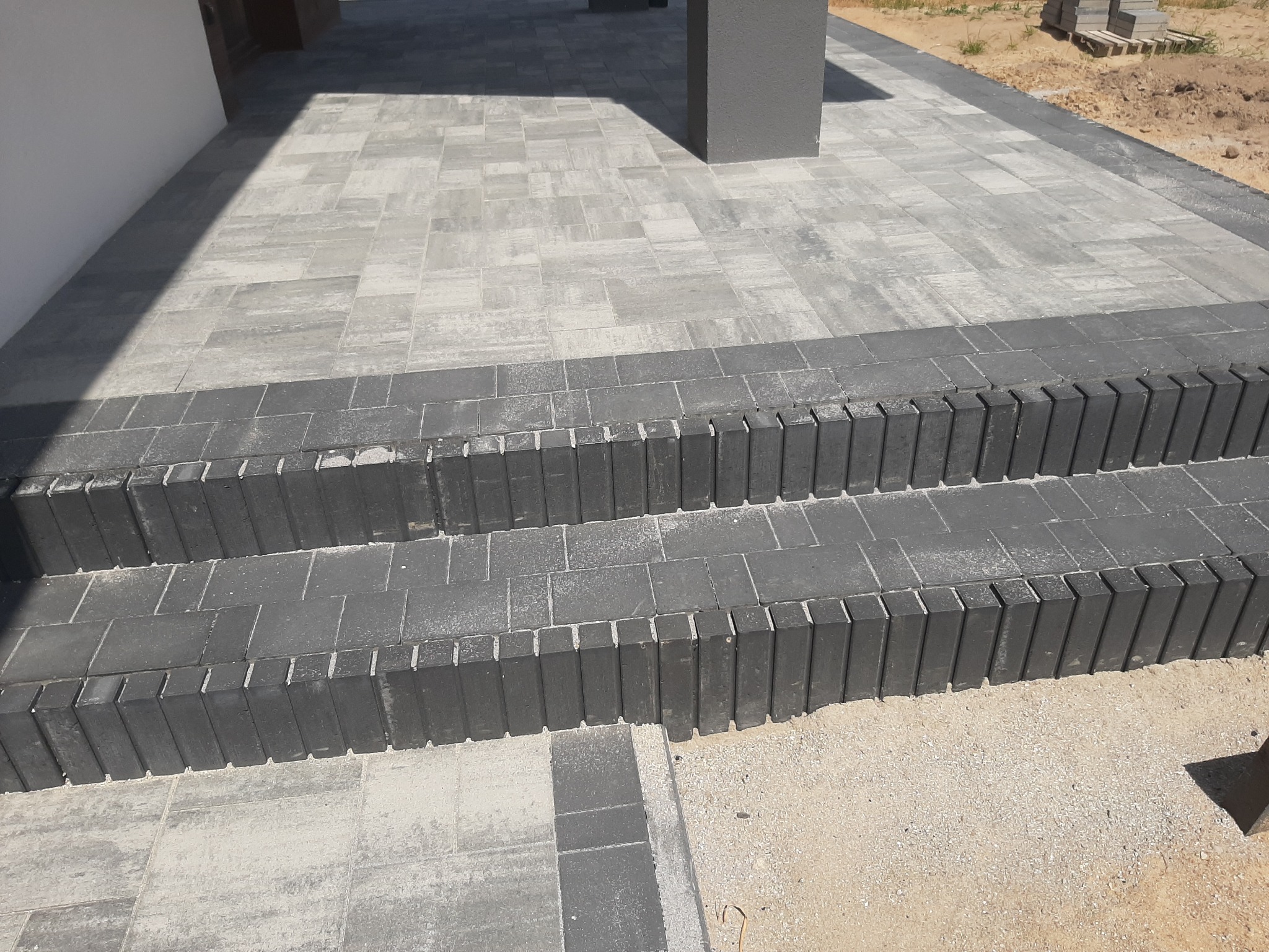 Schody wejściowe wyłożone szarą kostką brukową, z ciemnoszarymi obrzeżami i pionowymi elementami wykończeniowymi, prowadzące do domu z jasną elewacją.