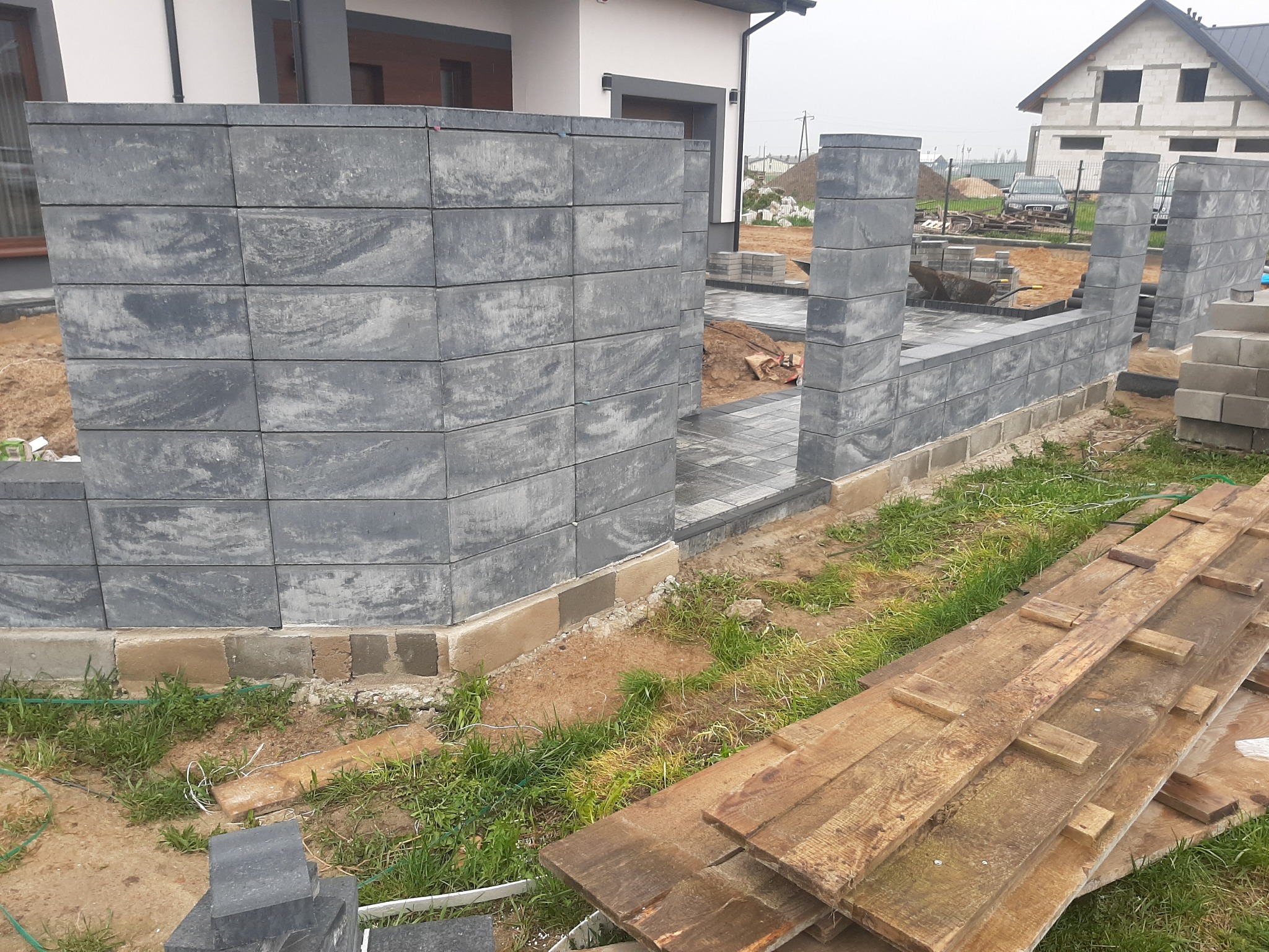 Budowa ogrodzenia z szarej kostki brukowej komorowej, widoczne fragmenty fundamentów, ułożona kostka prowadząca do bramy, deski budowlane w trawie.