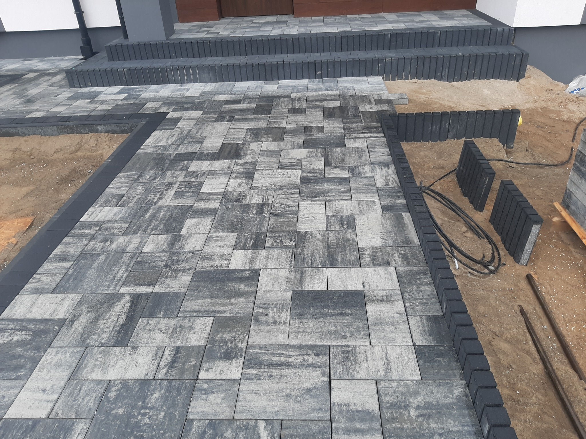 Niedokończona nawierzchnia z szaro-czarnej kostki brukowej o nieregularnym wzorze, z widocznymi fragmentami piasku i ułożonymi schodami z boku wejścia do budynku.